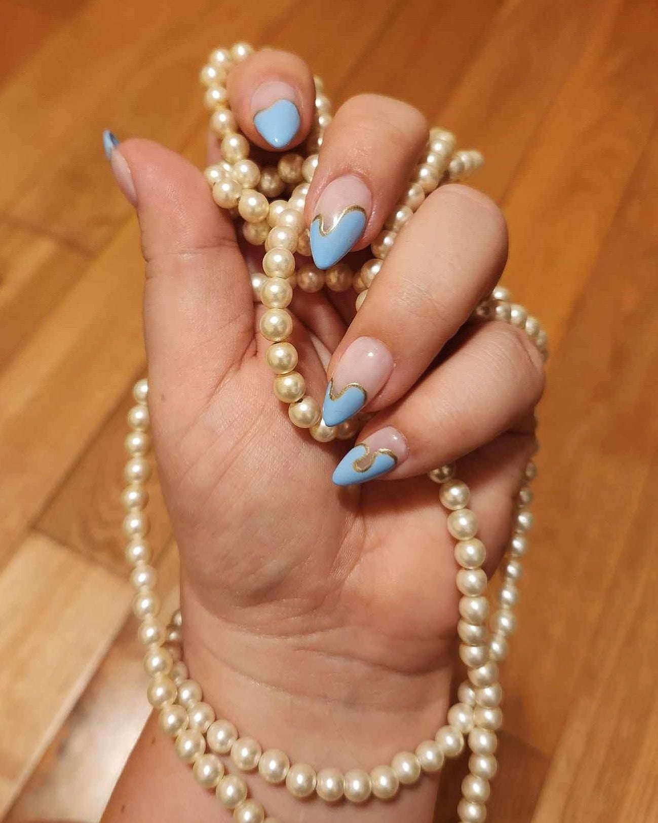 woman's hand holding a pearl necklace, showcasing a set of blue and gold swirl french tip press on nails